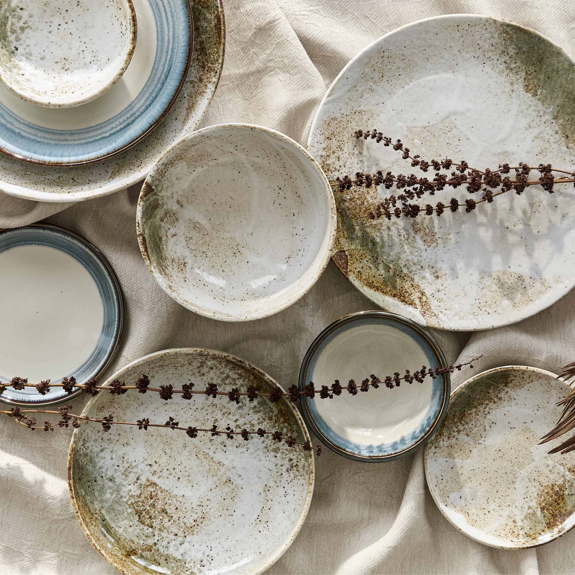 Aardewerken servies in natuurlijke tinten, met een combinatie van kommen, borden en schalen in witte, groenige en blauwe tinten, gedecoreerd met gedroogde takken op een beige tafelkleed.