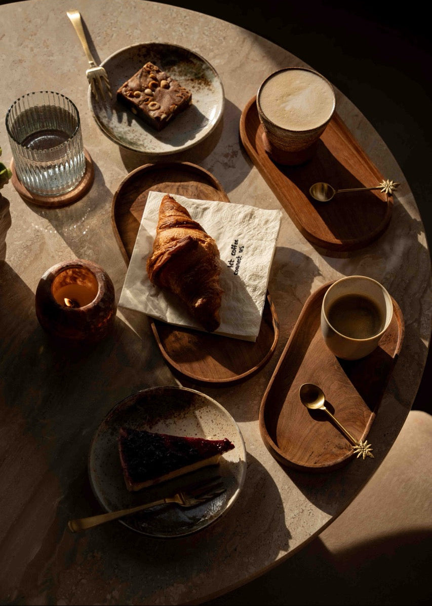 Warme brunchsetting met croissant op servet, mok latte en huisgemaakt taartje op keramiek en hout, onderdeel van de Café at Home tafelstyling collectie.