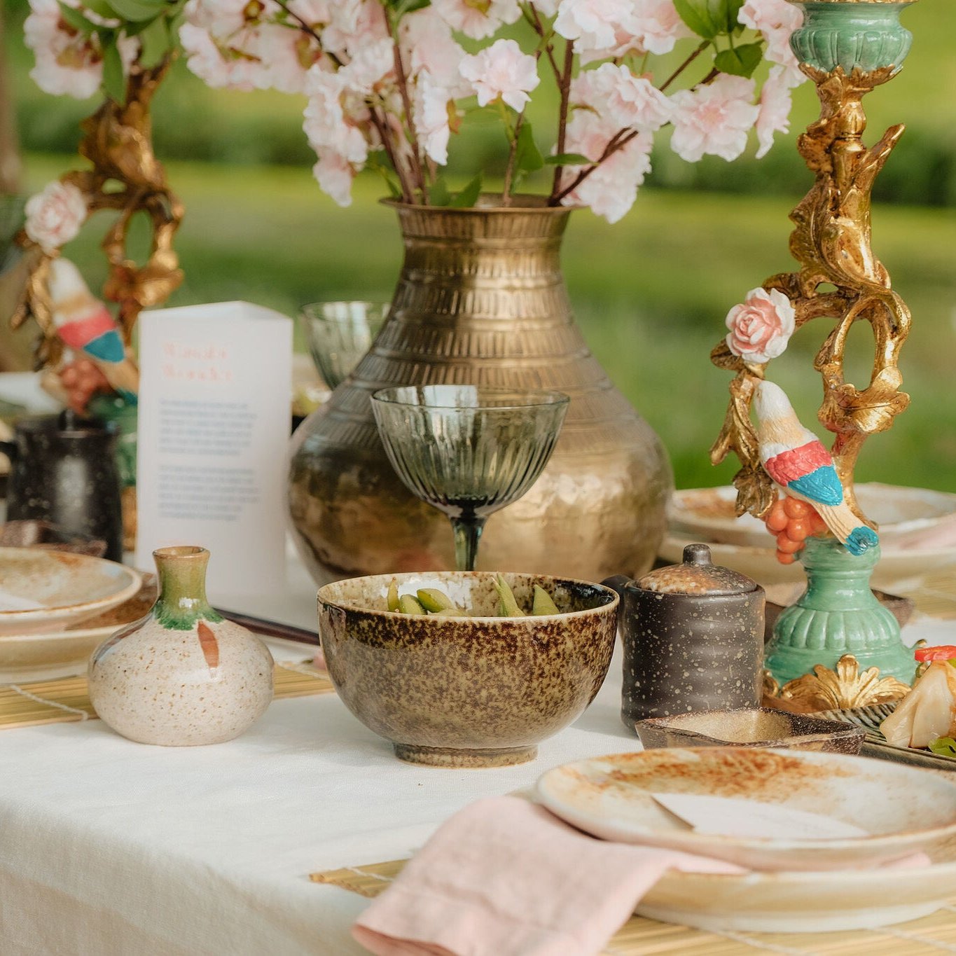 Elegant gedekte tafel met bronzen vaas gevuld met roze bloemen, kleurrijke kandelaar, groene wijnglazen, aardewerken servies, en een serveerschaal met dumplings.