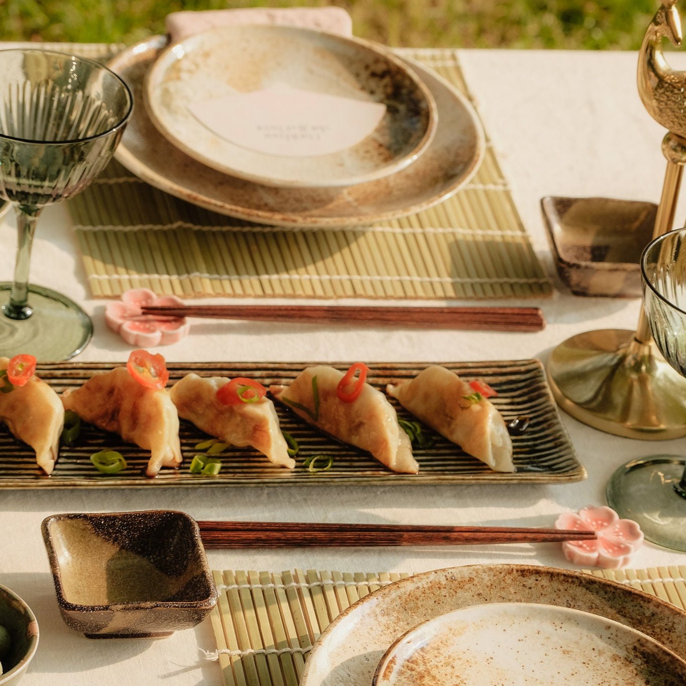 Gedekte tafel met groene wijnglazen, aardewerken servies, een gouden pauw kandelaar, en een serveerschaal met dumplings, gegarneerd met chili en lente-ui.