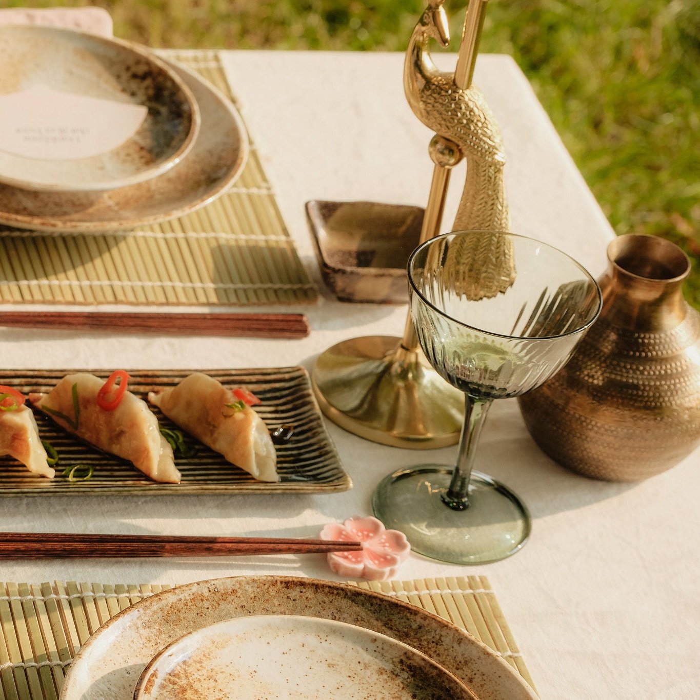 Gedekte tafel met groene wijnglazen, aardewerken servies, een gouden pauw kandelaar, en een serveerschaal met dumplings, gegarneerd met chili en lente-ui.
