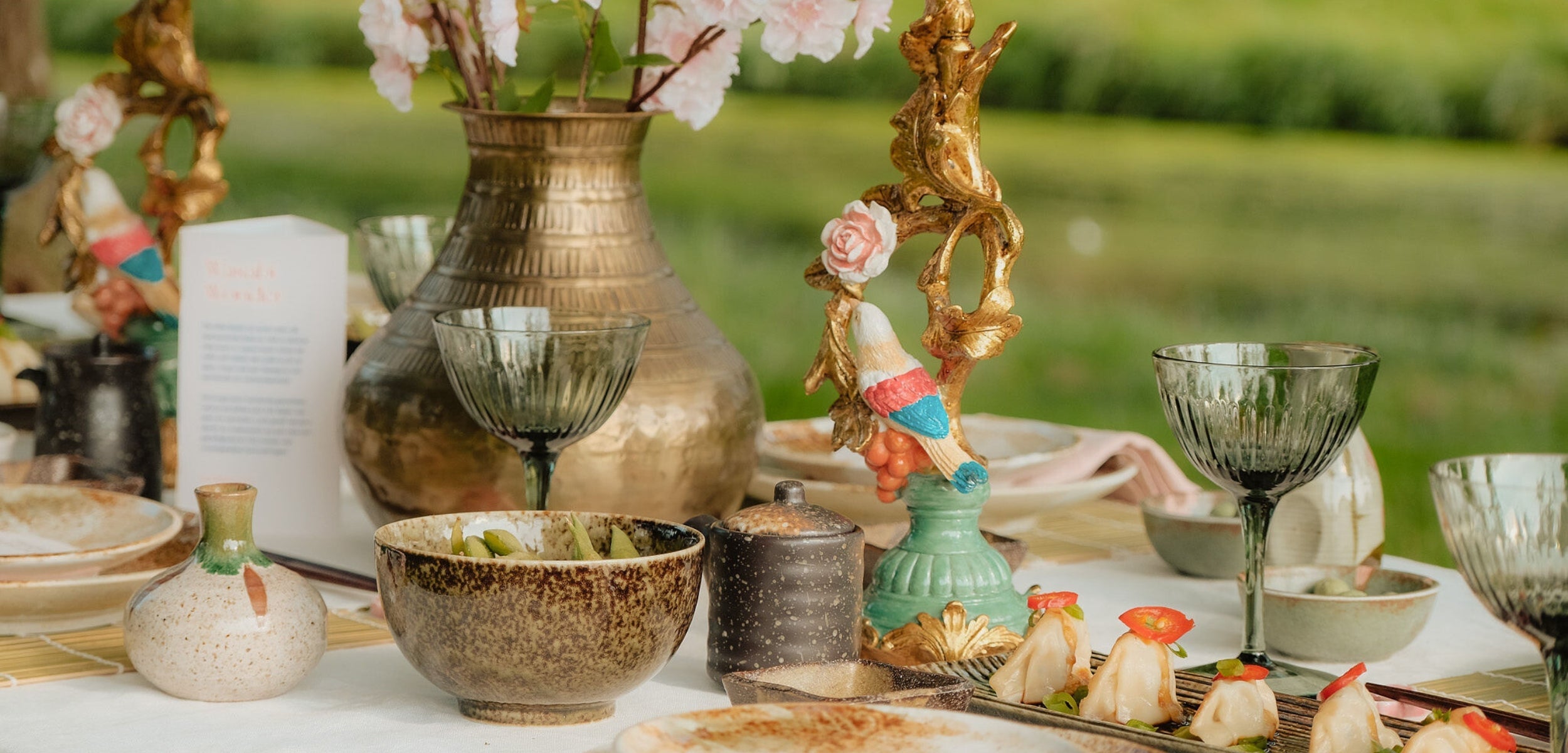 Gedekte tafel met een rustieke sfeer, centraal staat een bronzen vaas met roze bloesemtakken, omringd door gekleurde kandelaars, servies, glazen, en smaakvolle gerechten.