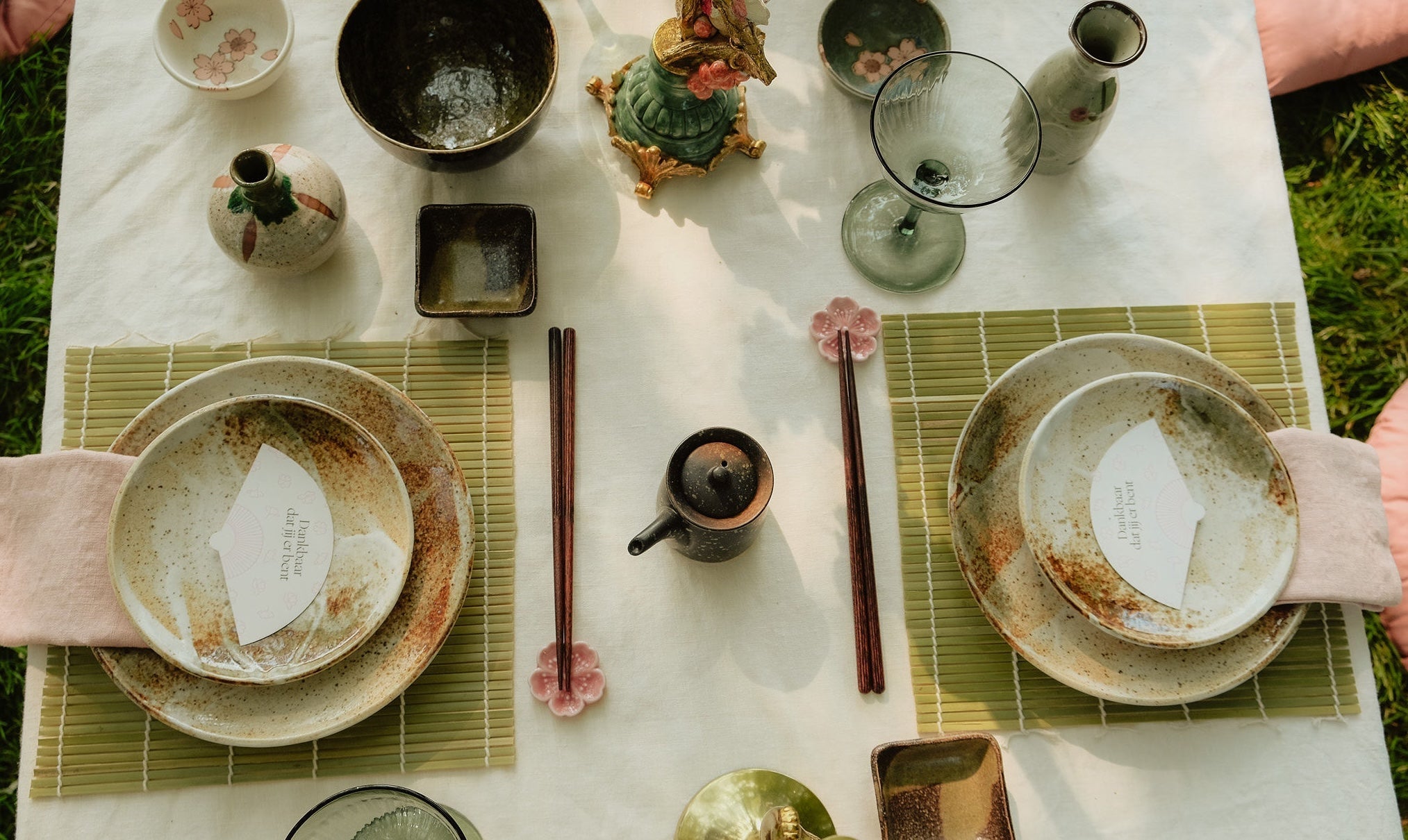 Bovenzicht van een gedekte tafel met rustieke borden, eetstokjes op bloesemhouders, een sake-fles en -kopje, en decoratieve elementen zoals een bloesemtak en een gouden kandelaar.