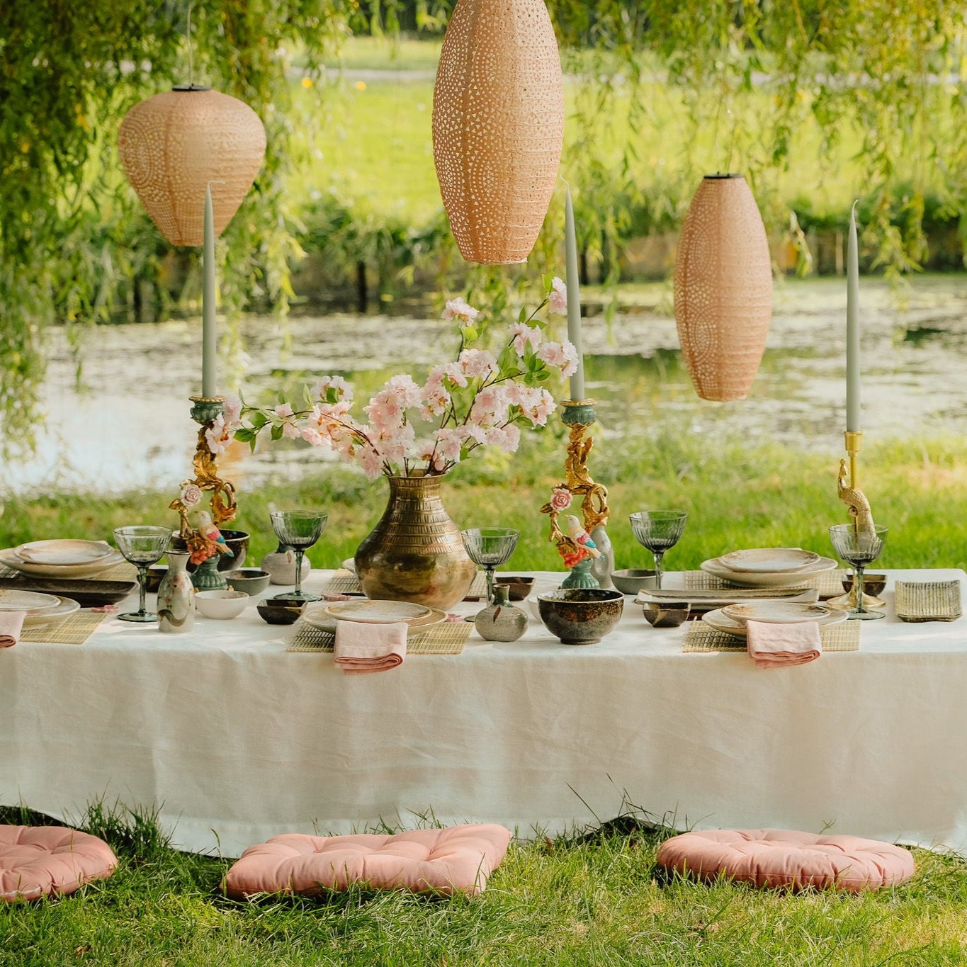 Sfeervolle picknicktafel aan de waterkant, gedecoreerd met bloemen, servies en kaarsen, onder hangende rieten lampionnen, met kussens op het gras.