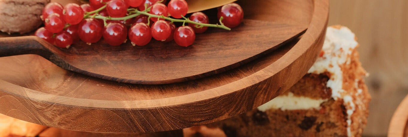 Close-up van een houten etagère met daarop een tros rode bessen en een stuk taart met roomvulling. Op de achtergrond zijn macarons en een ander stuk taart te zien.