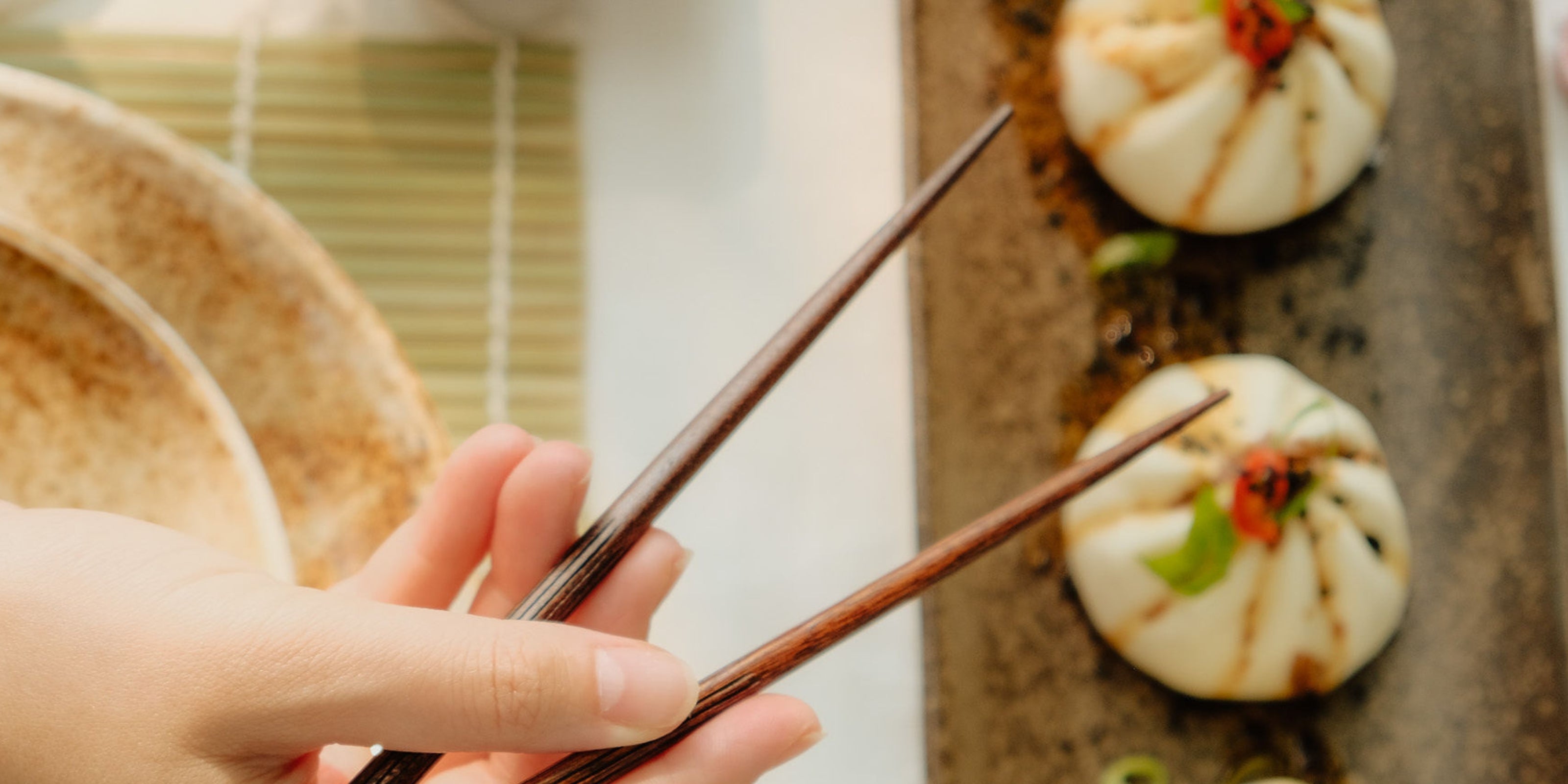 Close-up van een hand die eetstokjes vasthoudt, naast een bord met gestoomde broodjes, gedecoreerd met rode peper en lente-ui.