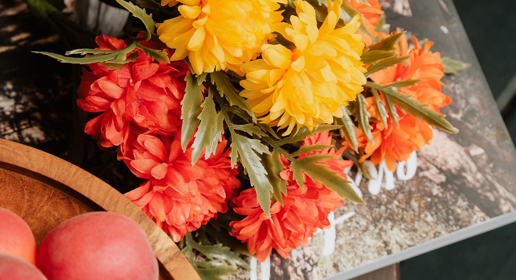 Close-up van een boeket kunstbloemen met felgele en oranje bloemen, geplaatst op een boek. Op de voorgrond is een houten schaal met roze perziken zichtbaar.