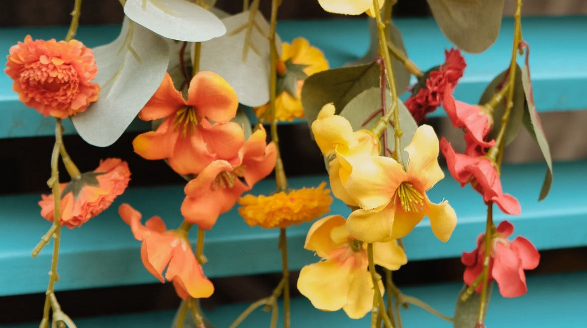 Close-up van een arrangement van kunstbloemen in oranje, gele en rode tinten, hangend tegen een turquoise houten achtergrond.