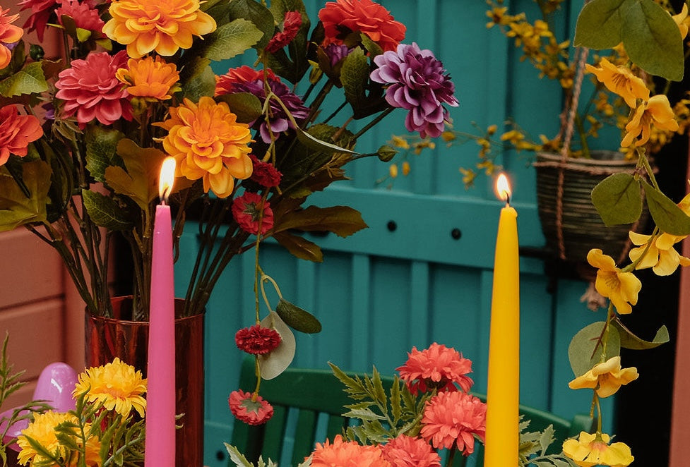 Close-up van een tafelschikking met kleurrijke kunstbloemen in een rode vaas, waaronder oranje, gele, paarse en rode bloemen. Op de voorgrond branden twee kaarsen, een roze en een gele, tegen een turquoise achtergrond.