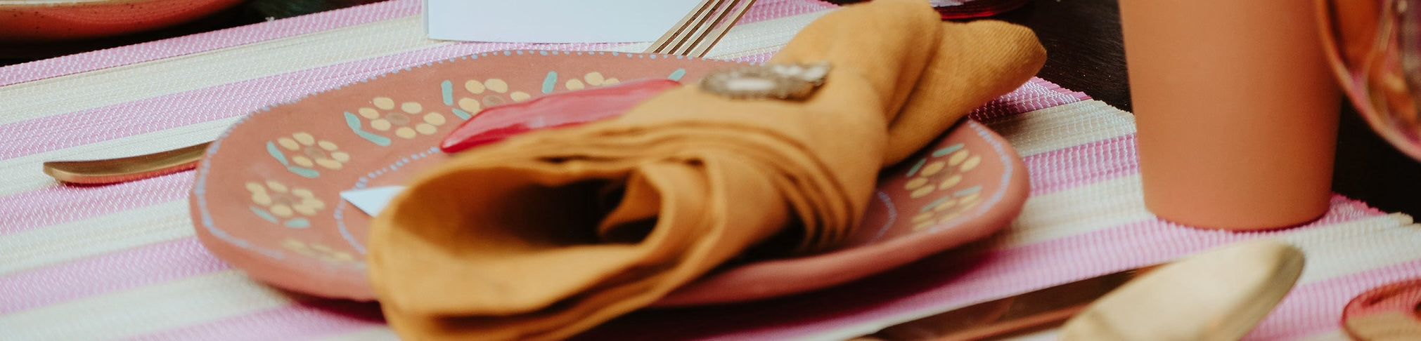 Close-up van een gedekte tafel met een roze bord met bloemmotief, een rode peper en een opgevouwen okergele servet. Het bord ligt op een roze-wit gestreept tafelkleed, met een gouden lepel en een terracotta beker op de achtergrond.