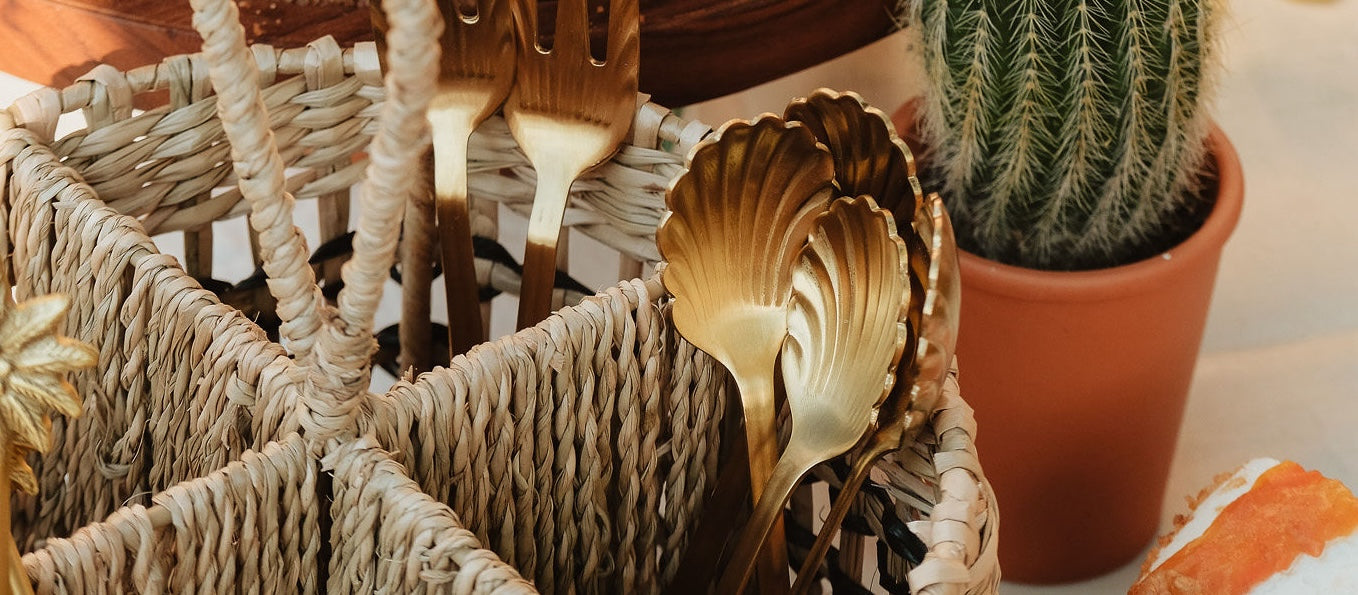 Close-up van een geweven mand met gouden bestek, waaronder vorken en lepels in de vorm van schelpen, naast een kleine cactus in een terracotta pot.