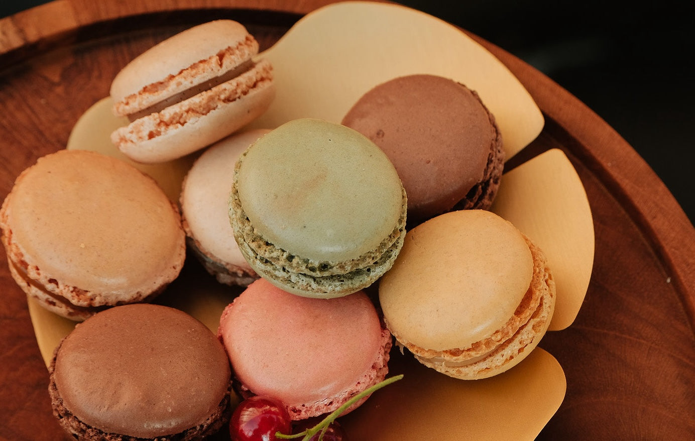 Close-up van een houten schaal met een selectie van kleurrijke macarons, waaronder groene, bruine, roze en beige macarons. Op de voorgrond ligt een tros rode bessen.