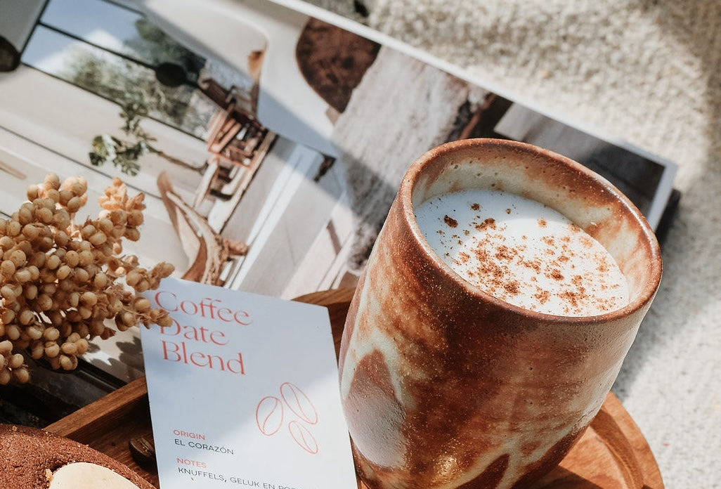 Close-up van een keramische mok met cappuccino bestrooid met kaneel, naast een kaart met de tekst 'Coffee Date Blend' en een droog bloemstuk. Op de achtergrond ligt een opengeslagen tijdschrift met interieurfoto's.