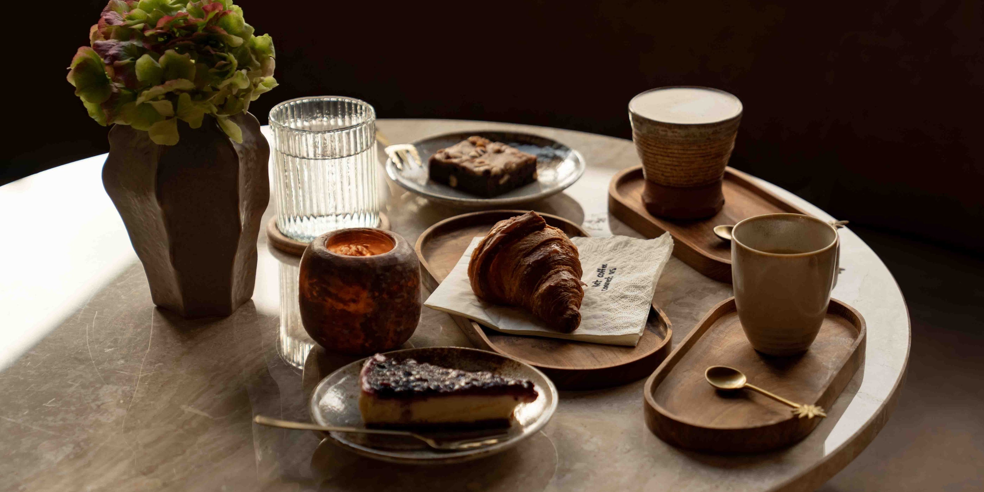 Warme café-aan-huis sfeer met koffie, croissant en gebak op houten serveerplanken – Café at Home collectie.