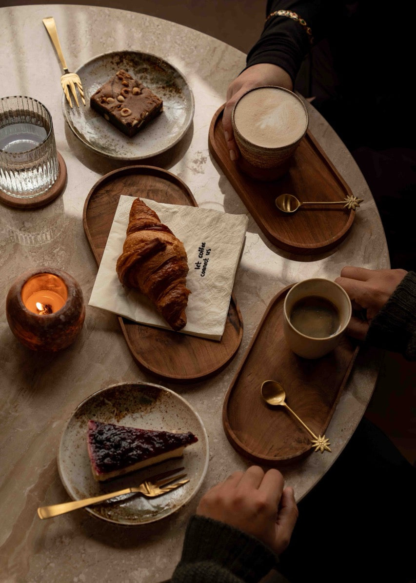 Sfeervolle koffietafel met croissant, cappuccino en gebak op houten serveerplanken uit de Café at Home collectie, gefotografeerd in warm en gezellig licht.