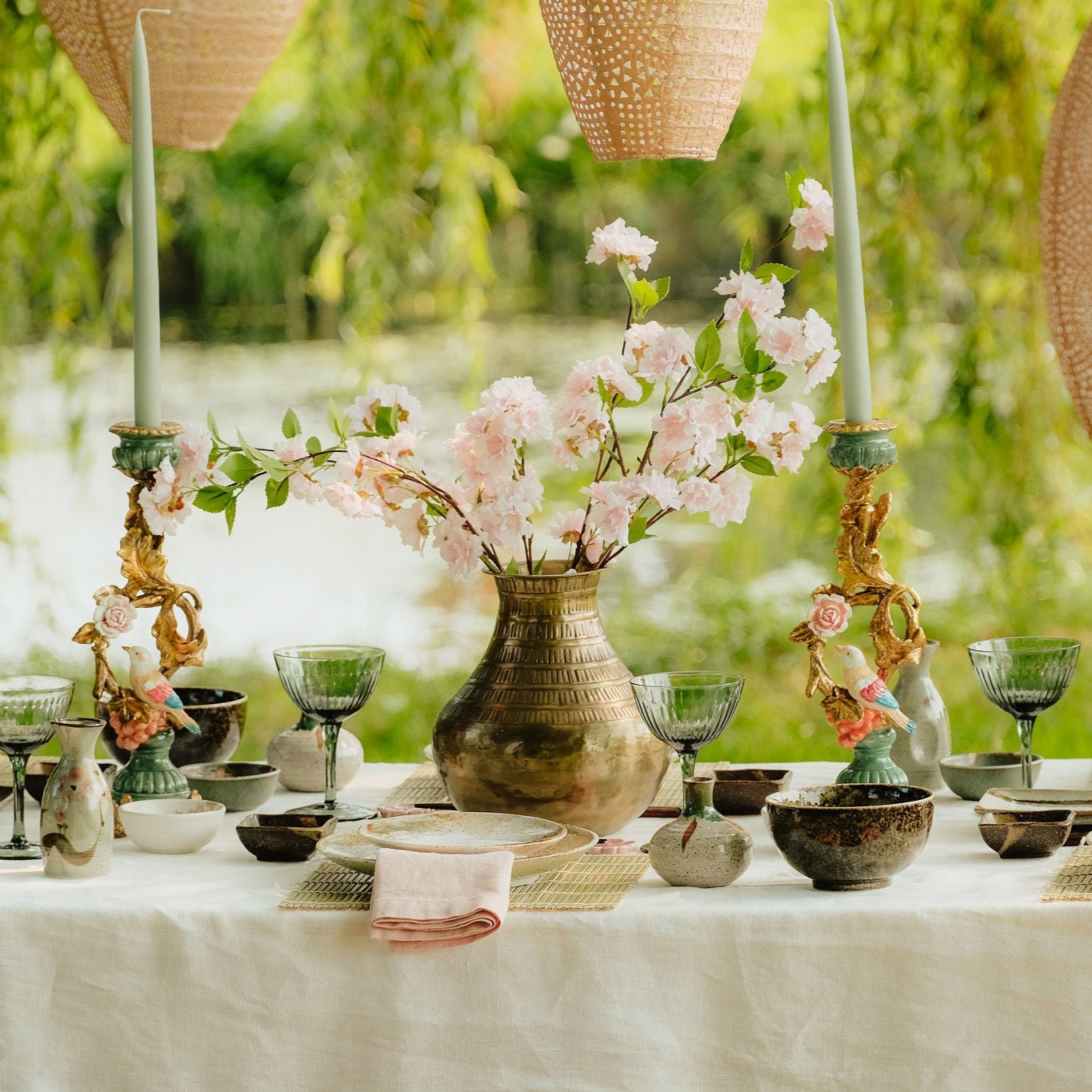 Sfeervolle picknicktafel aan de waterkant, gedecoreerd met bloemen, servies en kaarsen, onder hangende rieten lampionnen, met kussens op het gras.