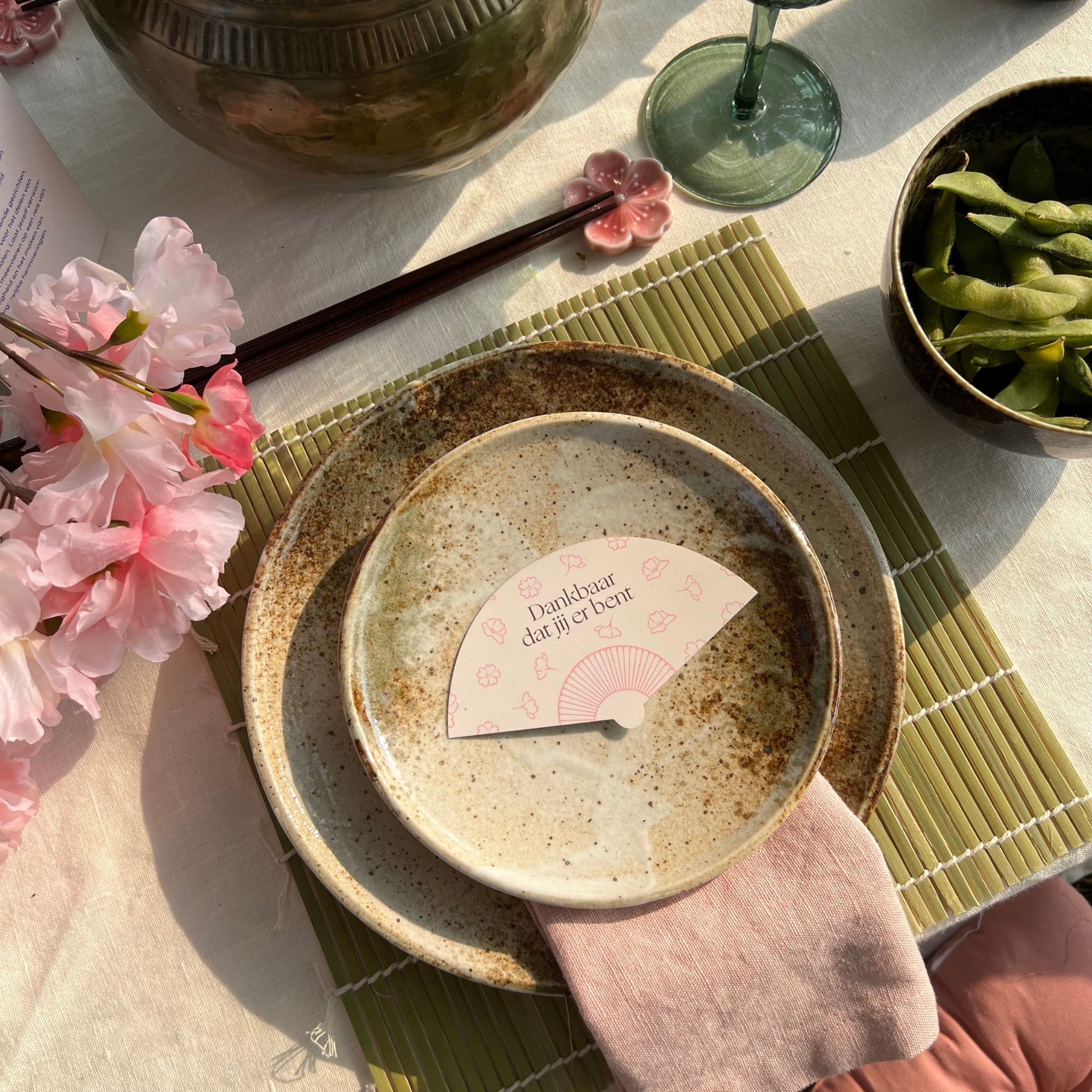 Bord met een roze linnen servet en een dankbaarheidskaartje in de vorm van een waaiertje, op een met zorg gedekte tafel met een Japanse sfeer. De tafel is versierd met roze bloesemtakken, eetstokjes, een bamboe placemat, en een schaaltje met edamamebonen.