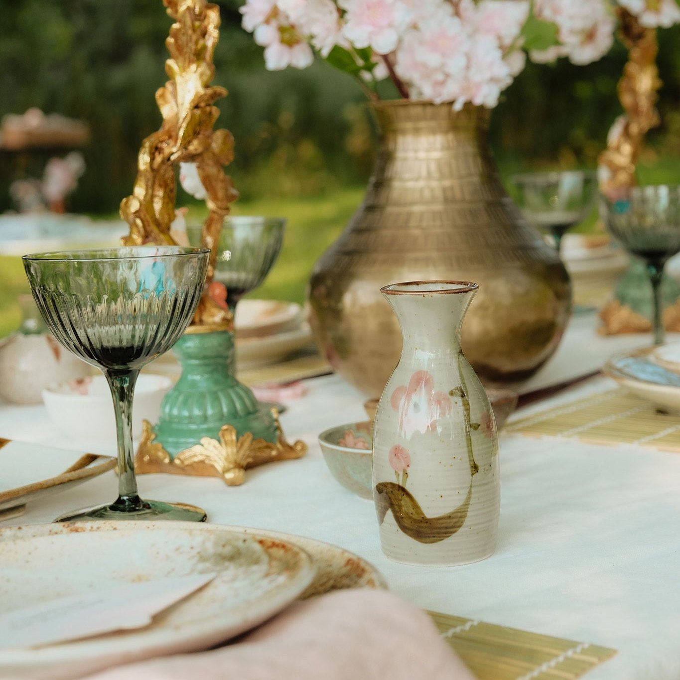 Gedekte tafel met een bronzen vaas gevuld met roze bloemen, groene wijnglazen, een decoratieve kandelaar, en een keramische sake fles met bloemmotief, op een wit tafelkleed.