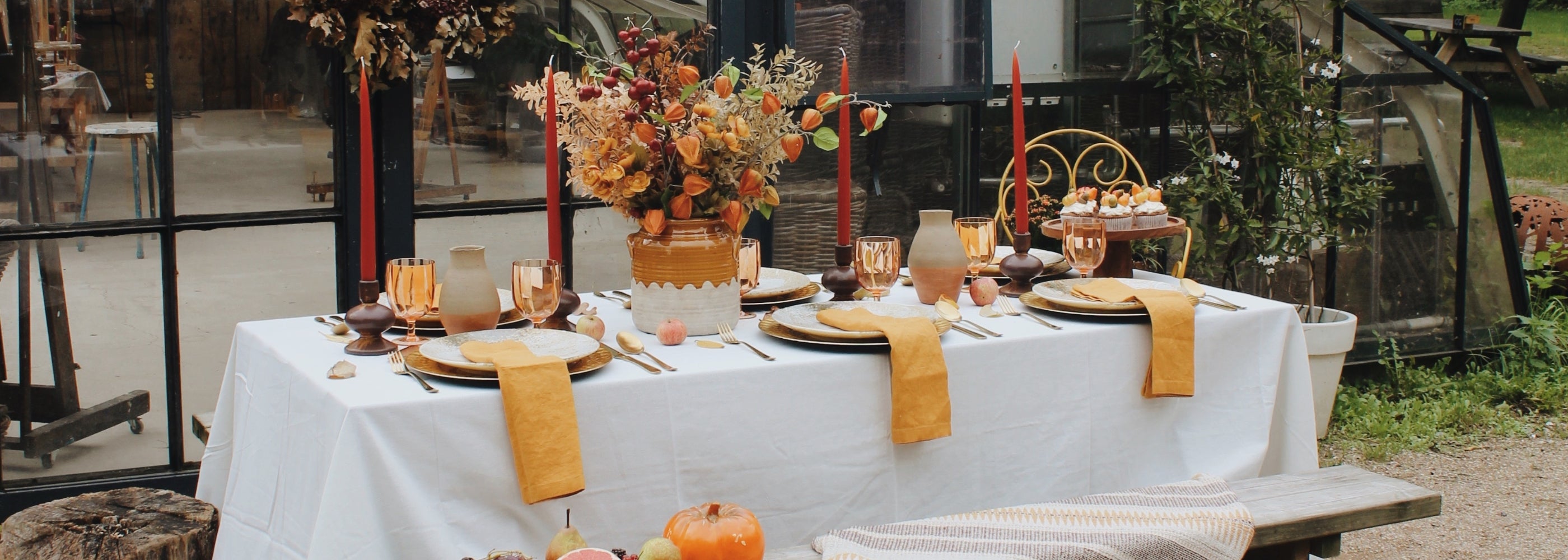 Buitentafel gedekt voor een sfeervol herfstdiner, compleet met amberkleurige glazen, gouden bestek, en een groot bloemstuk in warme oranje- en geelachtige tinten. De tafel, met witte linnen bekleding, is omgeven door seizoensdecoraties zoals pompoenen en appels. Lange rode kaarsen en een rustieke vaas met herfsttakken creëren een gezellige en uitnodigende ambiance voor een diner in de natuur.