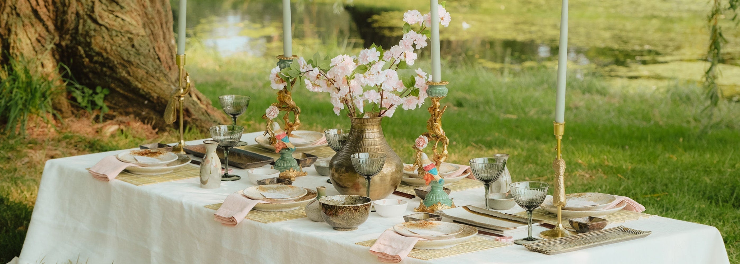 Elegante tafelsetting met een centraal geplaatste bronzen vaas gevuld met roze bloesemtakken, omringd door gedecoreerde kandelaars, borden, kommen, en glazen, gelegen aan een vijver.