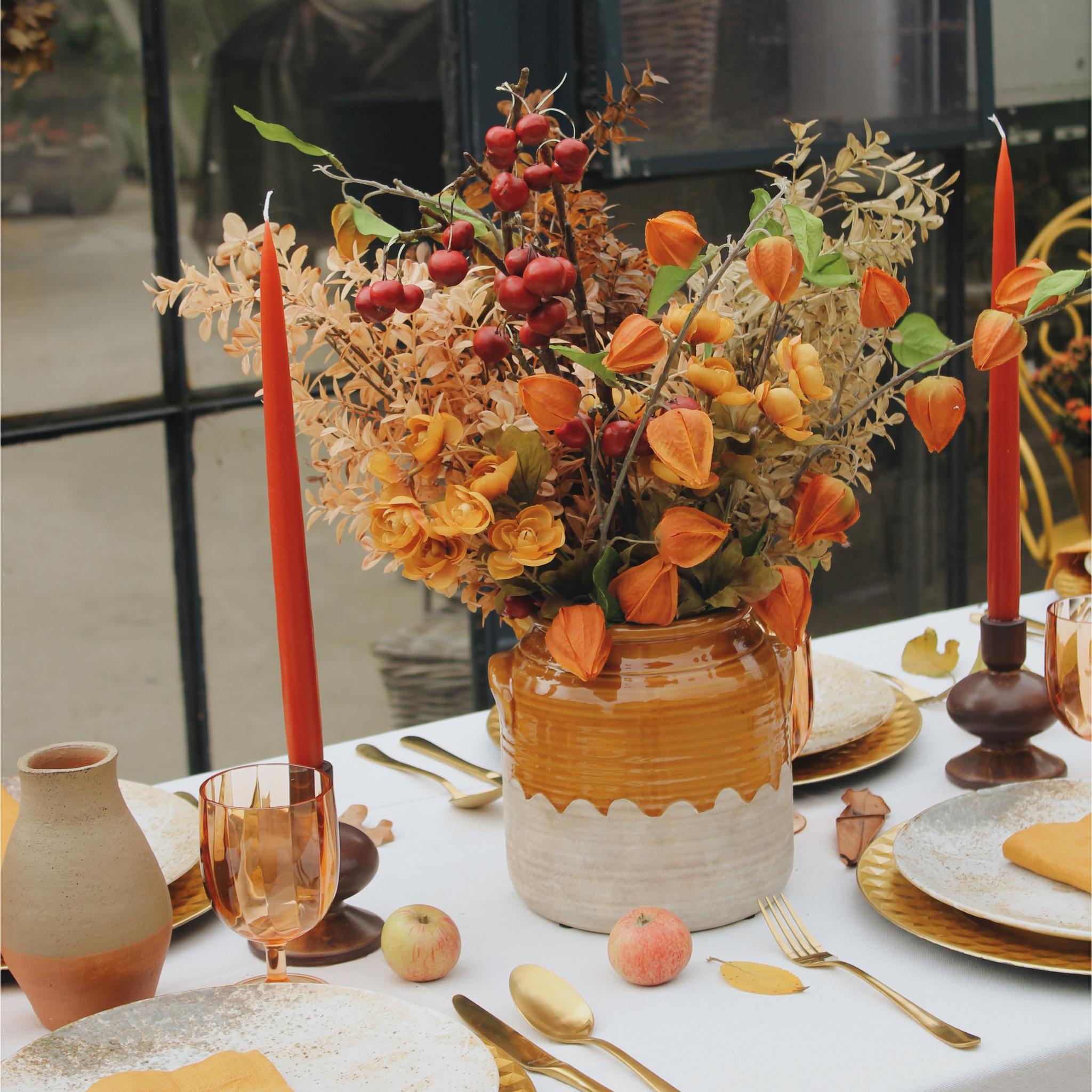 Detail van een herfsttafel met een prachtig bloemstuk in warme oranje- en goudtinten, geplaatst in een rustieke vaas op een wit tafellaken. De tafeldecoratie is aangevuld met gouden bestek, amberkleurige glazen, rode kaarsen en kleine decoratieve appels, wat de herfstige en gezellige sfeer benadrukt.