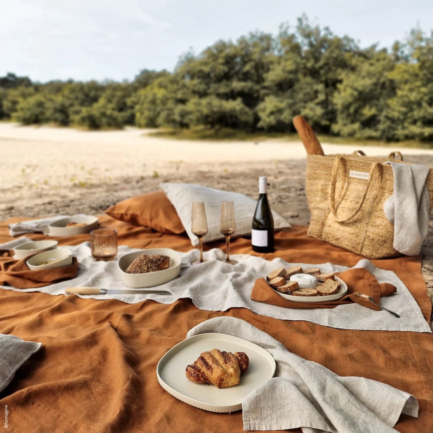 Picknick op een zandstrand met een mix van beige en oranje dekens, servies met croissants en brood, wijnglazen, een fles wijn, en een rieten tas, omgeven door een bosrijke omgeving.