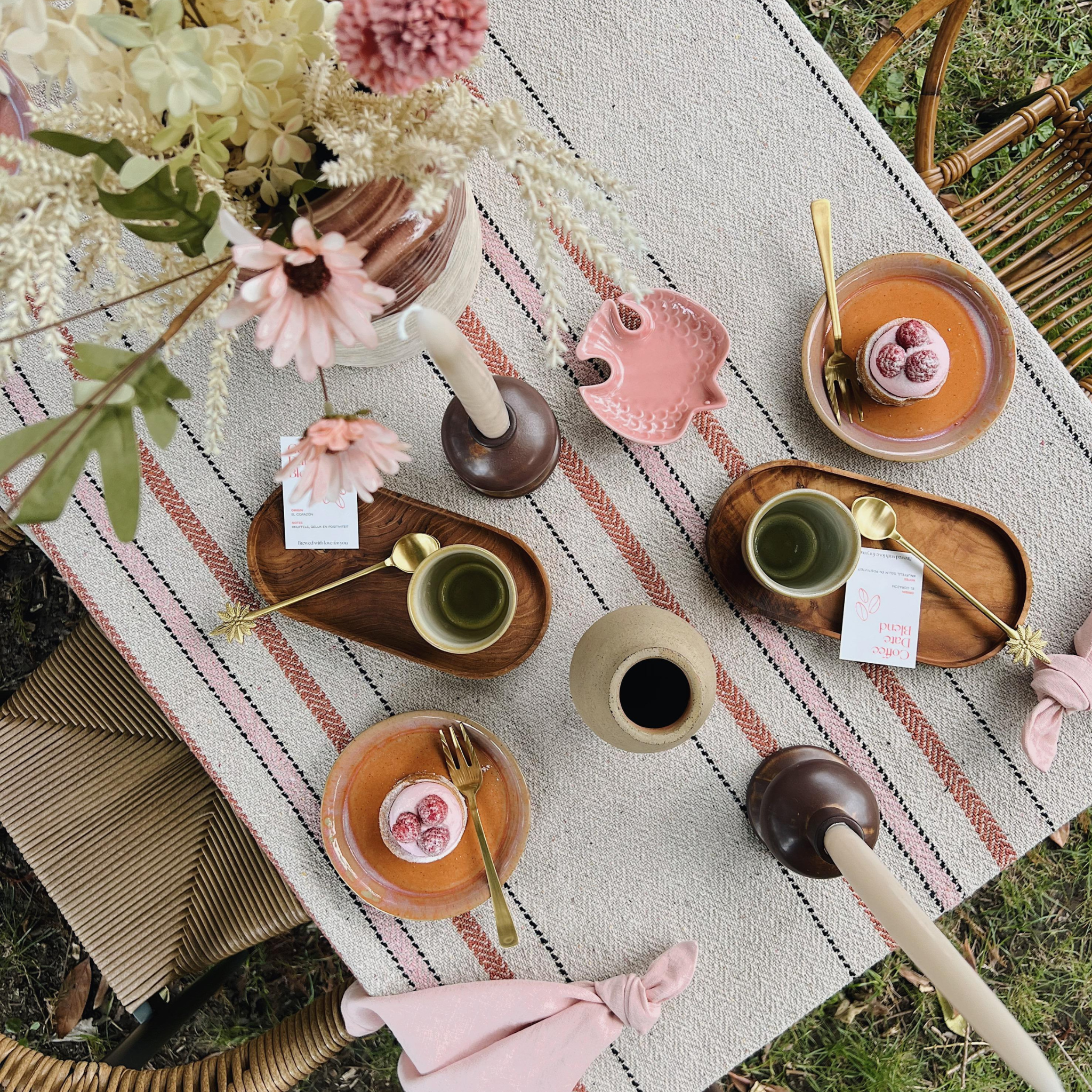 Bovenaanzicht van een sfeervol gedekte tafel in een natuurlijke omgeving. De tafeldecoratie bestaat uit een plaid met warme kleuren, bloemdecoratie, aardewerk servies, en gebakjes op borden met gouden bestek. De rustige en zachte tinten roze, beige en hout zorgen voor een uitnodigende en gezellige sfeer.