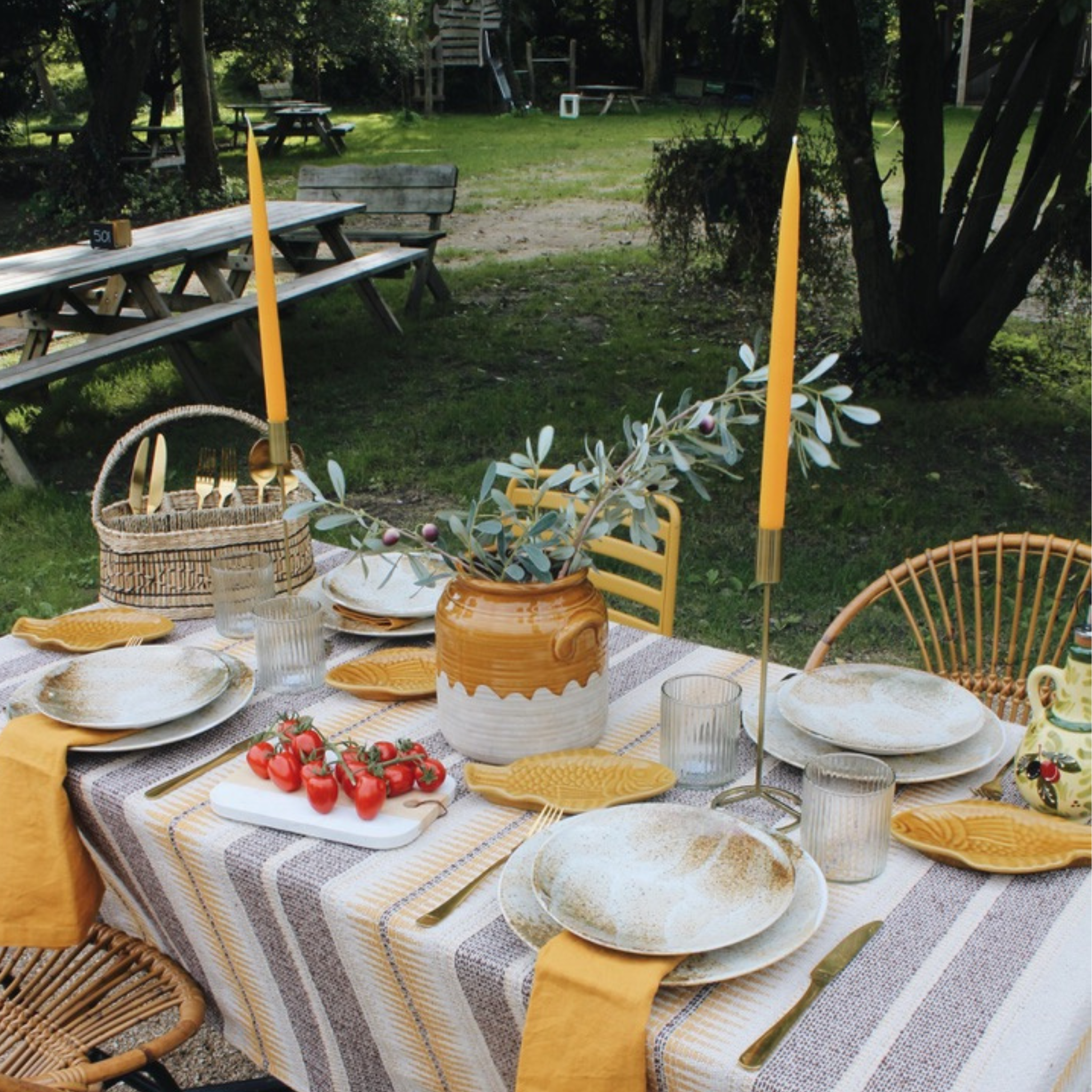 Buitentafel gedekt in mediterrane stijl met een tafelkleed in warme gele en beige tinten, aardewerk servies, en visvormige schalen. De tafel is versierd met gele kaarsen, een olijftak in een rustieke vaas, en een marmeren plank met trosjes rode tomaten. De setting straalt een warme, gezellige sfeer uit in een natuurlijke, groene omgeving.