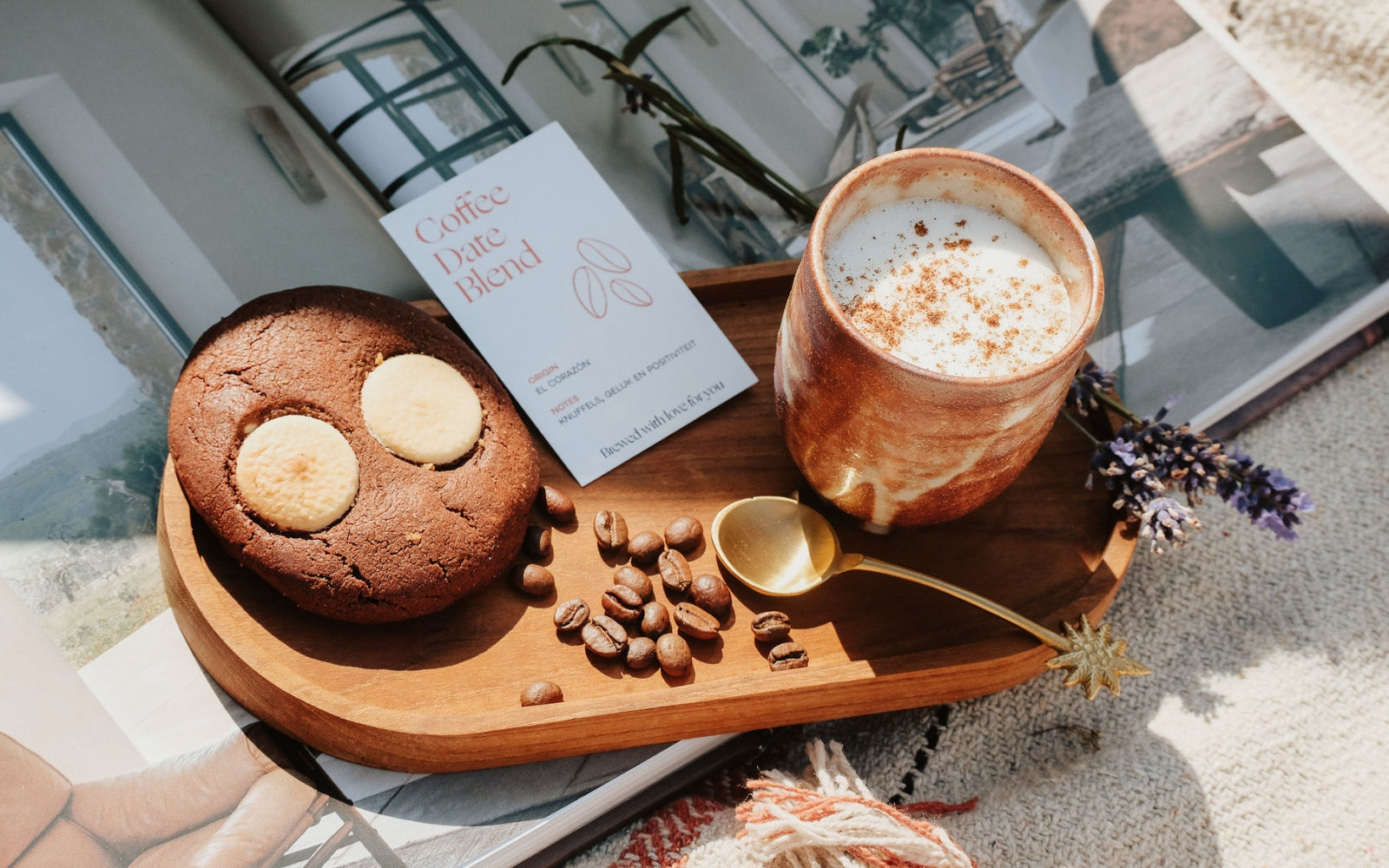 Een gezellige koffiemoment, inclusief een latte in een rustieke keramieken mok, een chocoladekoekje met witte chocolade, koffiebonen en een stijlvolle koffietag, gepresenteerd op een houten dienblad met een gouden lepel en lavendeltakjes in natuurlijk licht.