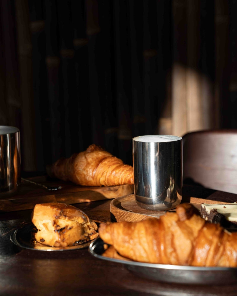 Warme ontbijttafel met stainless steel mokken, croissants en zachte ochtendzon – een sfeervolle, minimalistische setting vol rust en eenvoud.