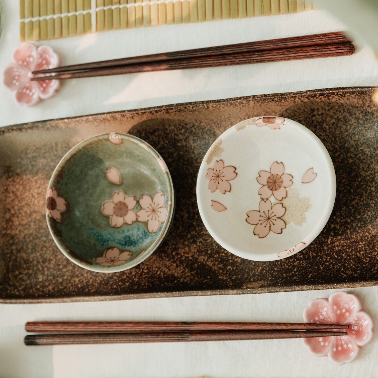 Tafelsetting met aardewerken serveerschaal, twee kleine kommen met sakura-bloesemmotief, houten eetstokjes en een groene wijnglas, op een witte tafellaken met bamboe placemat.