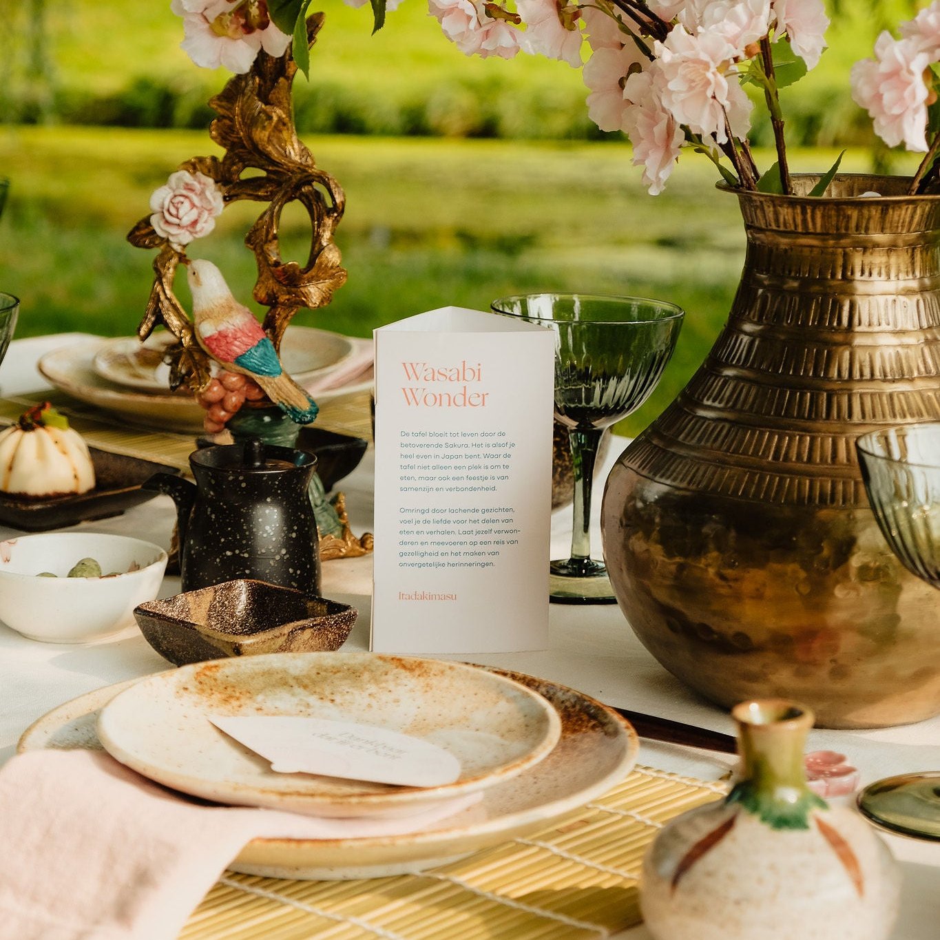 edekte tafel met een Japanse sfeer, inclusief een tafelkaartje met de tekst 'Wasabi Wonder'. De tafel is versierd met een grote vaas gevuld met roze bloesemtakken, goudkleurige kandelaars en elegant servies, wat een warme en uitnodigende uitstraling creëert.