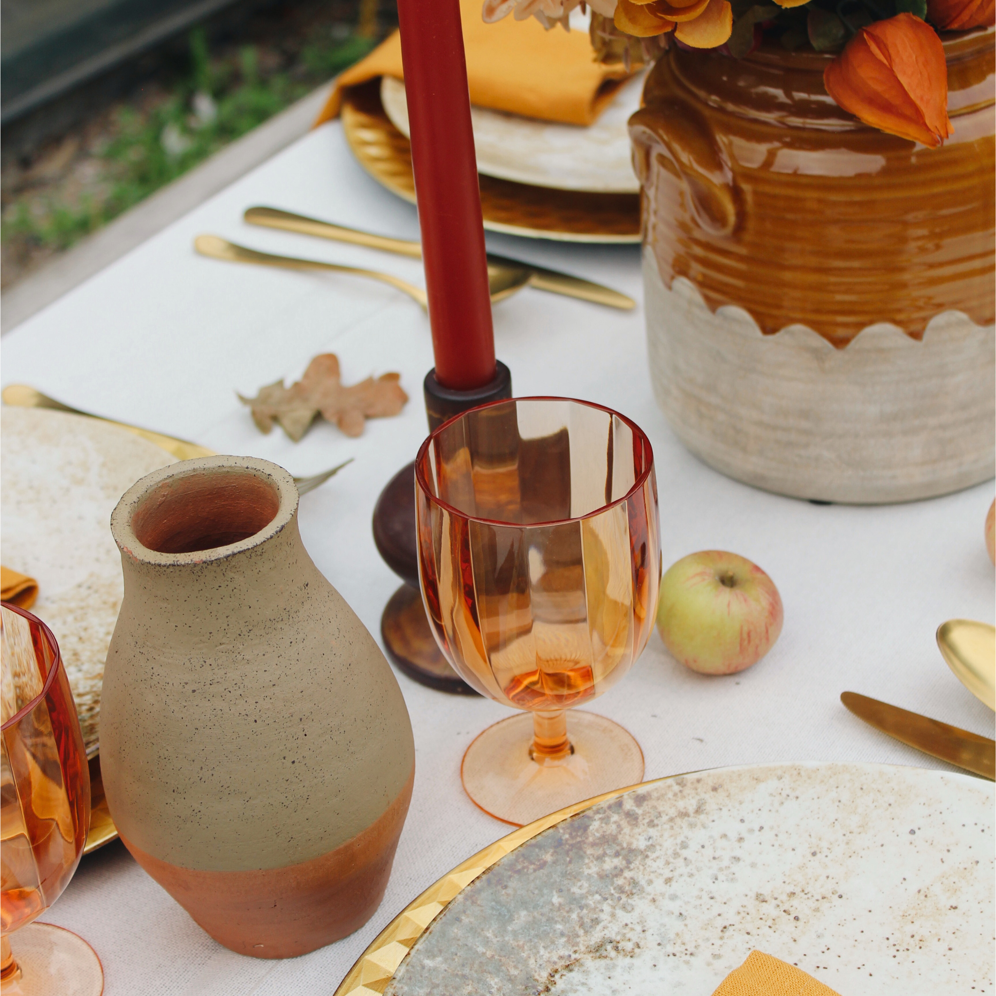 Detail van een herfstachtige tafeldecoratie met een rustieke terracotta vaas, amberkleurige wijnglazen, en een gedekte tafel in warme tinten. De tafel is versierd met een rode kaars, goudkleurig bestek, aardewerk borden en decoratieve appels. De warme herfstkleuren en natuurlijke elementen creëren een gezellige en uitnodigende sfeer.