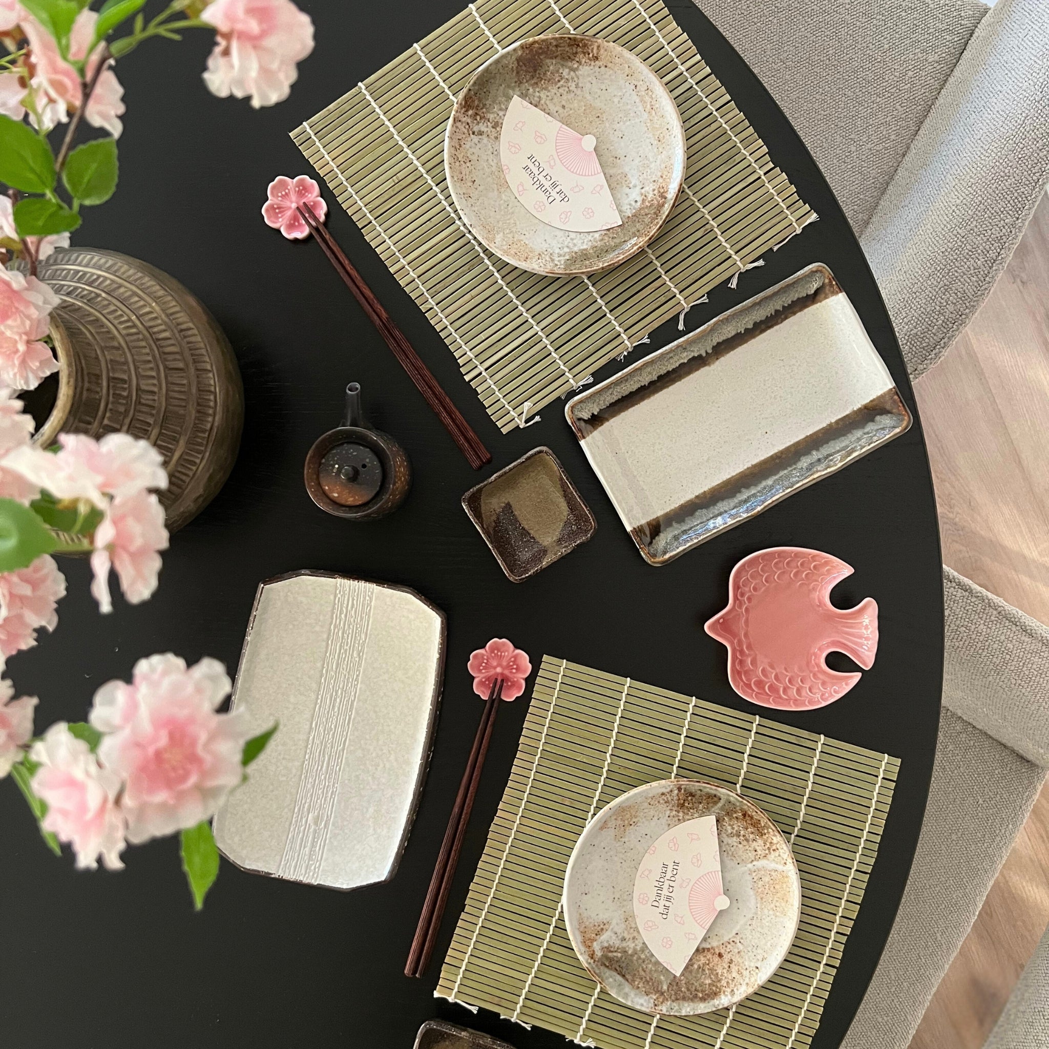 Sushi tafelsetting met bamboe placemats, Japanse keramieken servies, kersenbloesem eetstokjeshouders, en roze visvormige schotel, ideaal voor een thematische Sushi Date.